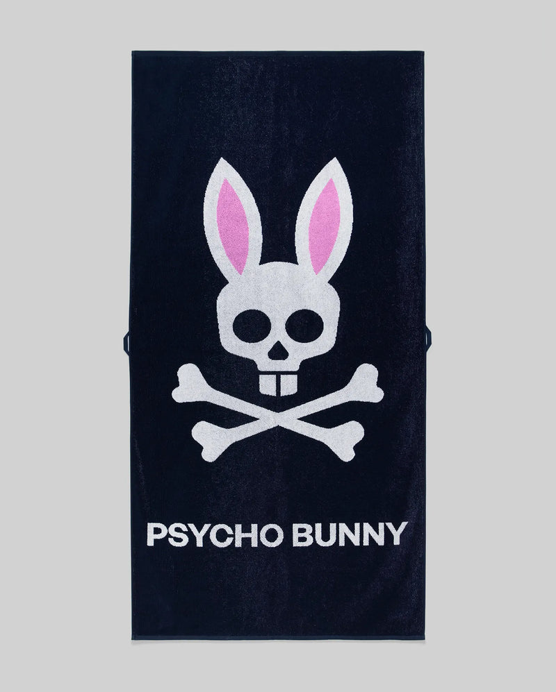 The Psycho Bunny MENS CLASSIC BUNNY TOWEL (MO1700004) is a dark towel with a white skull and crossbones topped by pink bunny ears above bold "PSYCHO BUNNY" lettering.