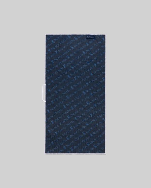 A rectangular navy MENS MICROFIBER TOWEL - MO1700007 by Psycho Bunny, featuring a subtle diagonal repeating logo pattern, is laid flat on a light gray background with a small fabric tag in the upper right corner.