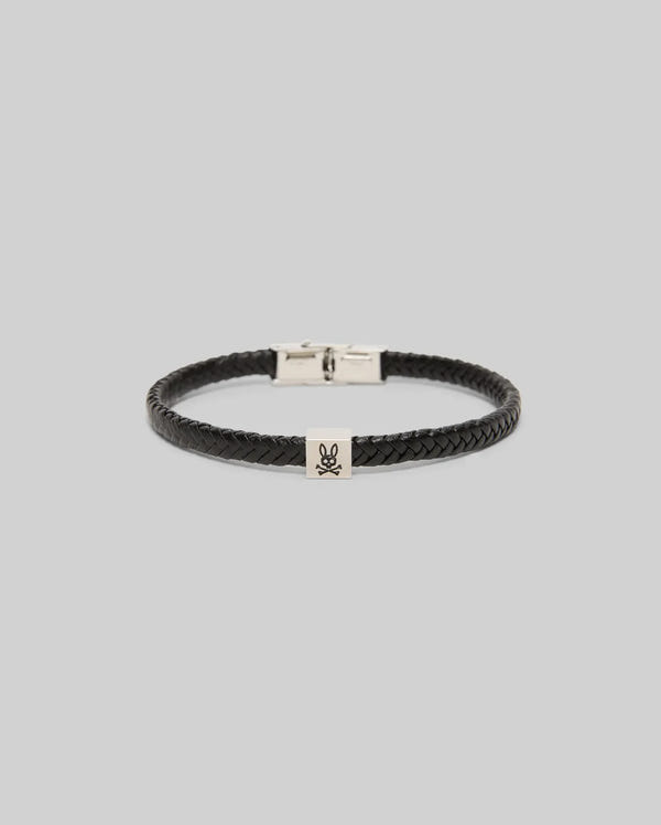 The Psycho Bunny MENS LEATHER BRAIDED BRACELET - MO1700020 features black braided leather, a silver rectangular hinged clasp, and a small engraved skull and crossbones charm, displayed on a light gray background.