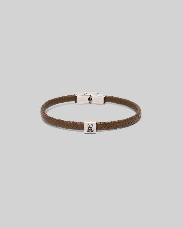 The MENS LEATHER BRAIDED BRACELET - MO1700020 by Psycho Bunny features brown braided leather, bunny-engraved hardware, a silver clasp, and a small silver charm with a black bunny skull and crossbones on a gray background.