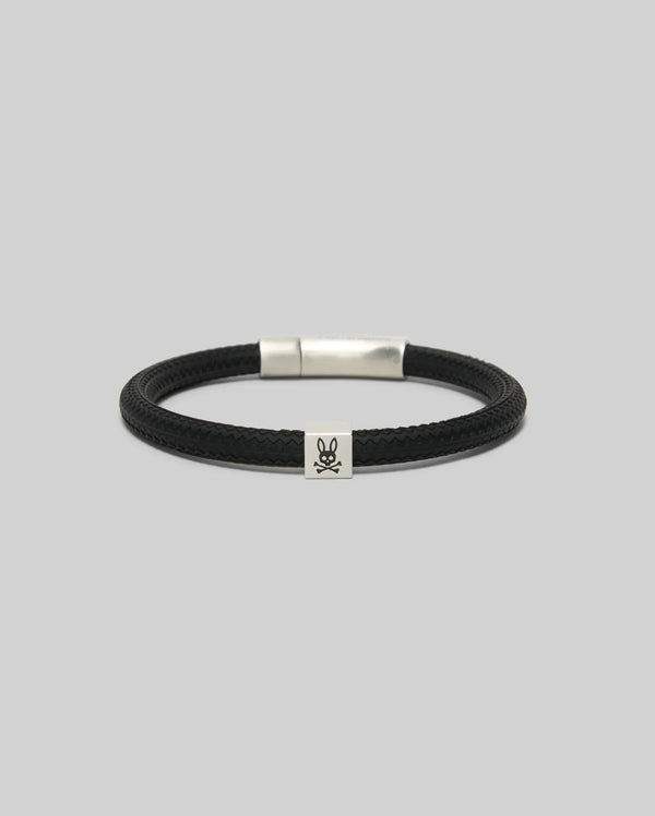The Psycho Bunny MENS SILICONE BRACELET - MO1700025 features a black braided design, a rectangular silver clasp, and a small silver skull and crossbones charm, all displayed on a plain grey background.