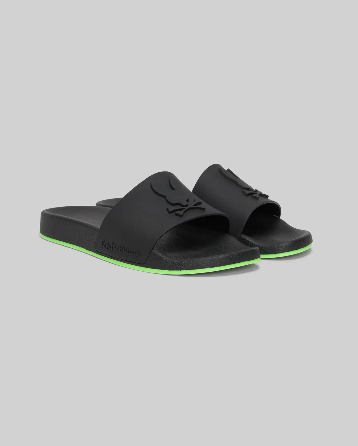 The Psycho Bunny MENS CONTRAST SOLE SLIDES (MO2400007) are black slide sandals with a raised logo on the strap, a green stripe on the sole, and a molded footbed for comfort, shown against a plain gray background.