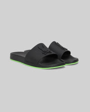 The Psycho Bunny MENS CONTRAST SOLE SLIDES (MO2400007) are black slide sandals with a raised logo on the strap, a green stripe on the sole, and a molded footbed for comfort, shown against a plain gray background.