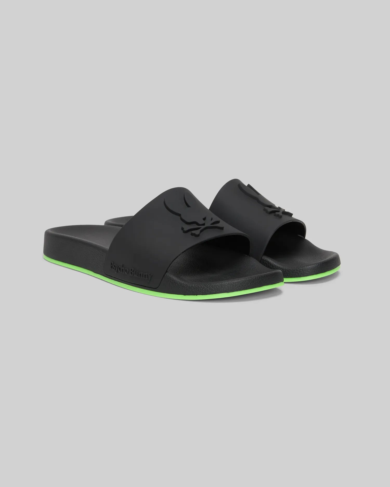 The Psycho Bunny MENS CONTRAST SOLE SLIDES (MO2400007) are black slide sandals with a raised logo on the strap, a green stripe on the sole, and a molded footbed for comfort, shown against a plain gray background.