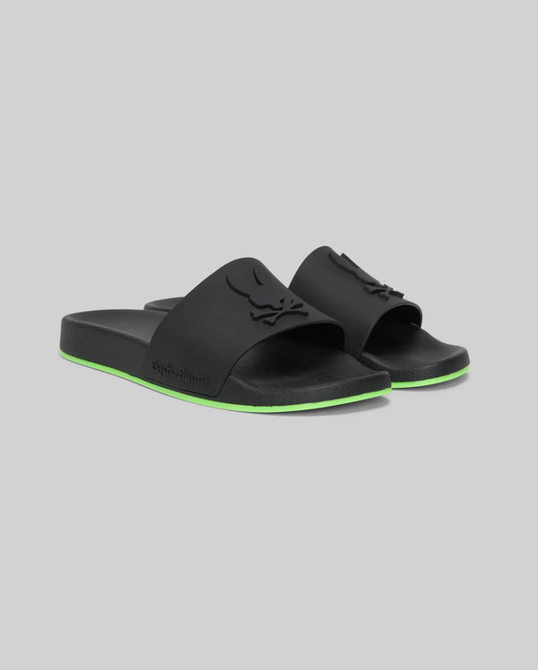 The Psycho Bunny MENS CONTRAST SOLE SLIDES (MO2400007) are black slide sandals with a raised logo on the strap, a green stripe on the sole, and a molded footbed for comfort, shown against a plain gray background.