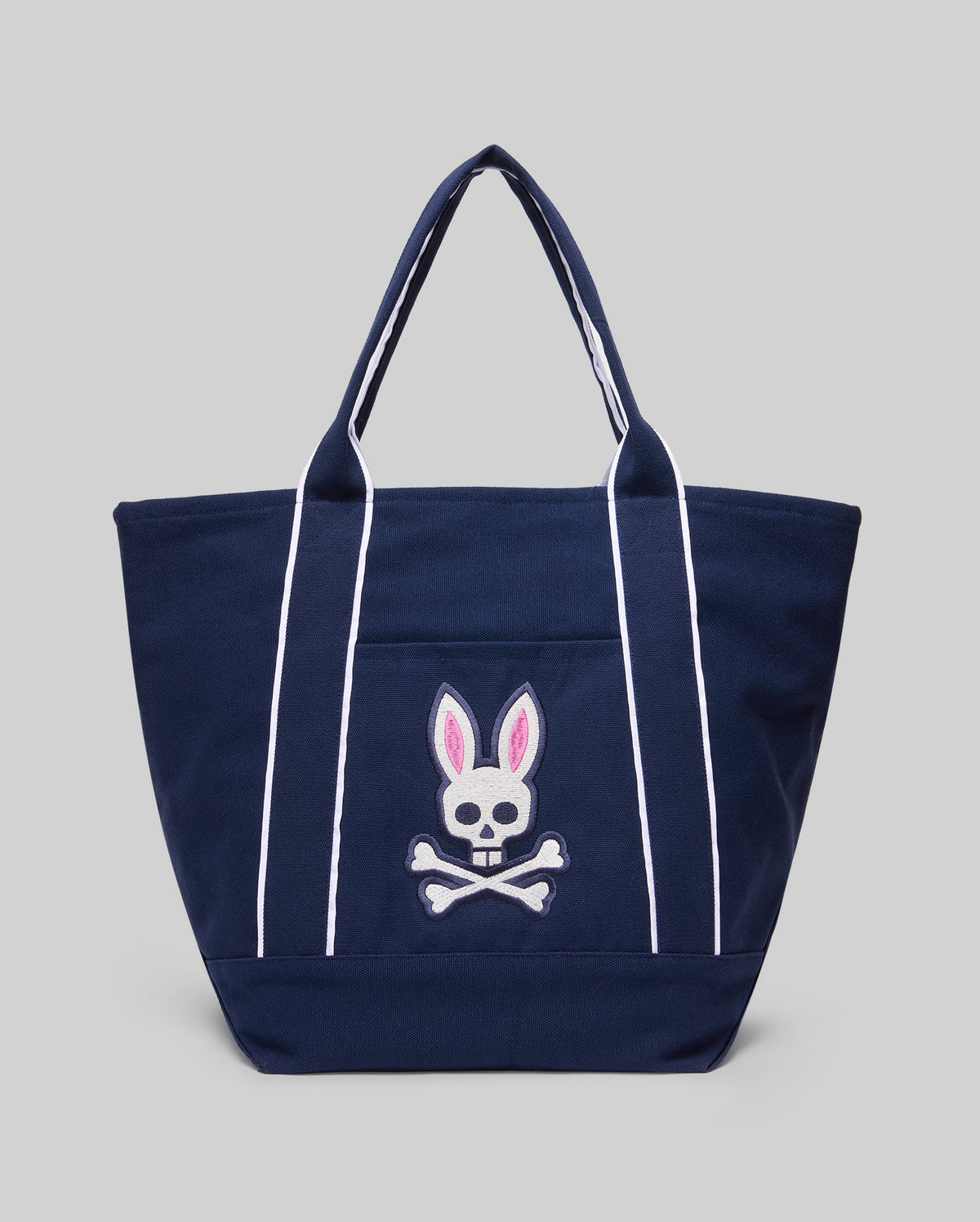 The Psycho Bunny MENS CANVAS TOTE BAG - MO2400016 is a navy blue tote with white handles and stripes, plus an embroidered skull and crossbones with pink bunny ears. Made for durability, it’s a stylish match for premium athleisure. Light gray background.