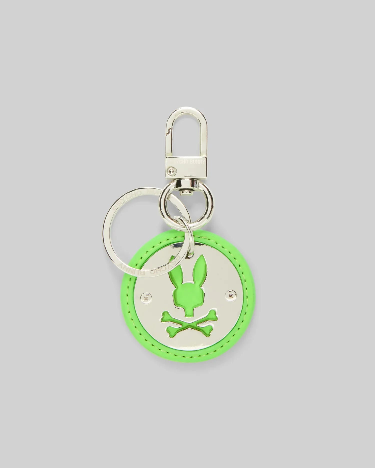 The DISC KEYCHAIN - MO2400020 by Psycho Bunny is a round neon green disc keychain featuring a silver bunny head with crossed bones cutout, attached to a polished metal keyring and lanyard clasp, displayed on a plain gray background.