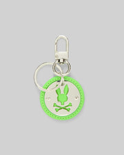 The DISC KEYCHAIN - MO2400020 by Psycho Bunny is a round neon green disc keychain featuring a silver bunny head with crossed bones cutout, attached to a polished metal keyring and lanyard clasp, displayed on a plain gray background.