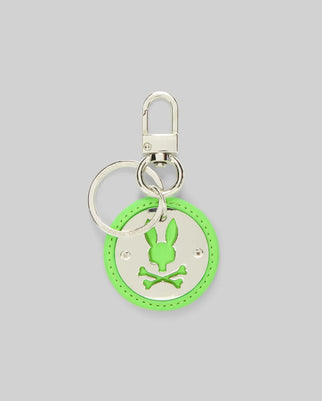 The DISC KEYCHAIN - MO2400020 by Psycho Bunny is a round neon green disc keychain featuring a silver bunny head with crossed bones cutout, attached to a polished metal keyring and lanyard clasp, displayed on a plain gray background.