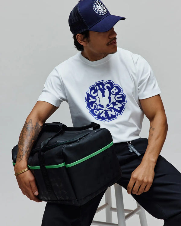 White classic fit jersey t-shirt with a large blue Psycho Bunny graphic, navy trucker cap with white embroidered logo, black relaxed pants, and a Black Gym Bag featuring green accents and a subtle all-over logo pattern.
