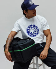 White classic fit jersey t-shirt with a large blue Psycho Bunny graphic, navy trucker cap with white embroidered logo, black relaxed pants, and a Black Gym Bag featuring green accents and a subtle all-over logo pattern.