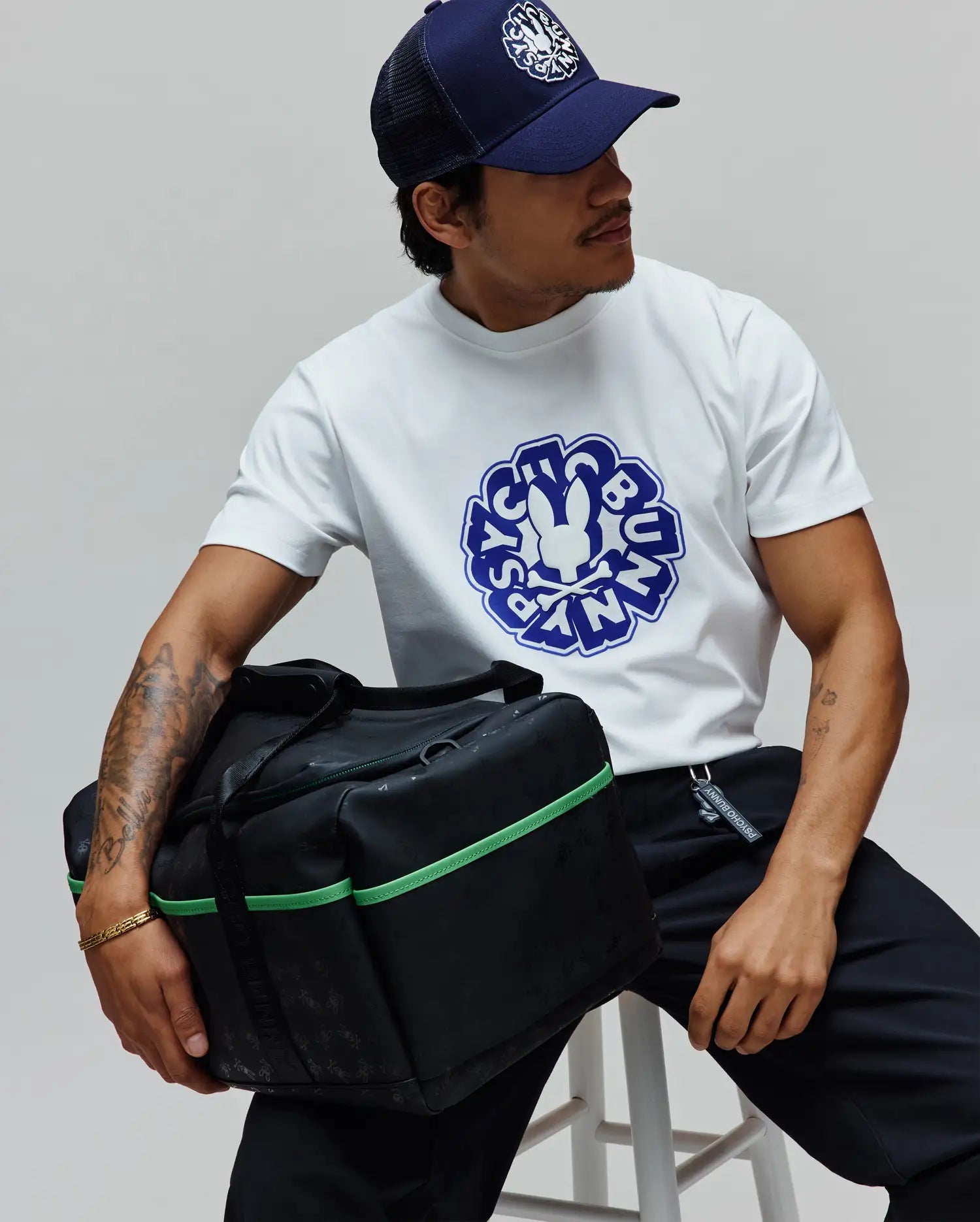 White classic fit jersey t-shirt with a large blue Psycho Bunny graphic, navy trucker cap with white embroidered logo, black relaxed pants, and a Black Gym Bag featuring green accents and a subtle all-over logo pattern.