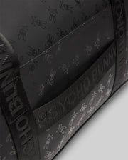 Black Gym Bag featuring an all-over tonal dark grey Psycho Bunny skull and crossbones logo pattern, complemented by woven webbing straps with repeated Psycho Bunny text branding.