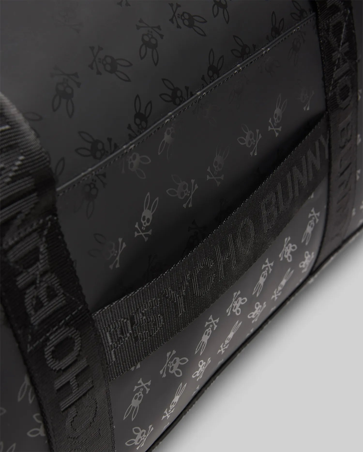 Black Gym Bag featuring an all-over tonal dark grey Psycho Bunny skull and crossbones logo pattern, complemented by woven webbing straps with repeated Psycho Bunny text branding.