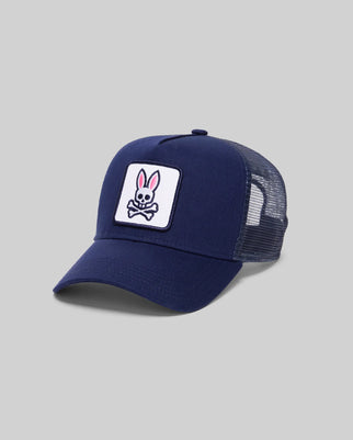 The Psycho Bunny MENS TRUCKER CAP - MO2600001 is a navy blue cap with a mesh back and a front patch showing a pink and white bunny above crossed bones, creating a playful skull and crossbones look, set against a plain gray background.