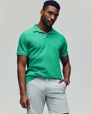 A man wears the Psycho Bunny MENS JAXON SOFT PIQUE CAMP COLLAR POLO - MT0100006 in green with light gray shorts, standing against a plain background, one hand in his pocket and the other relaxed by his side.