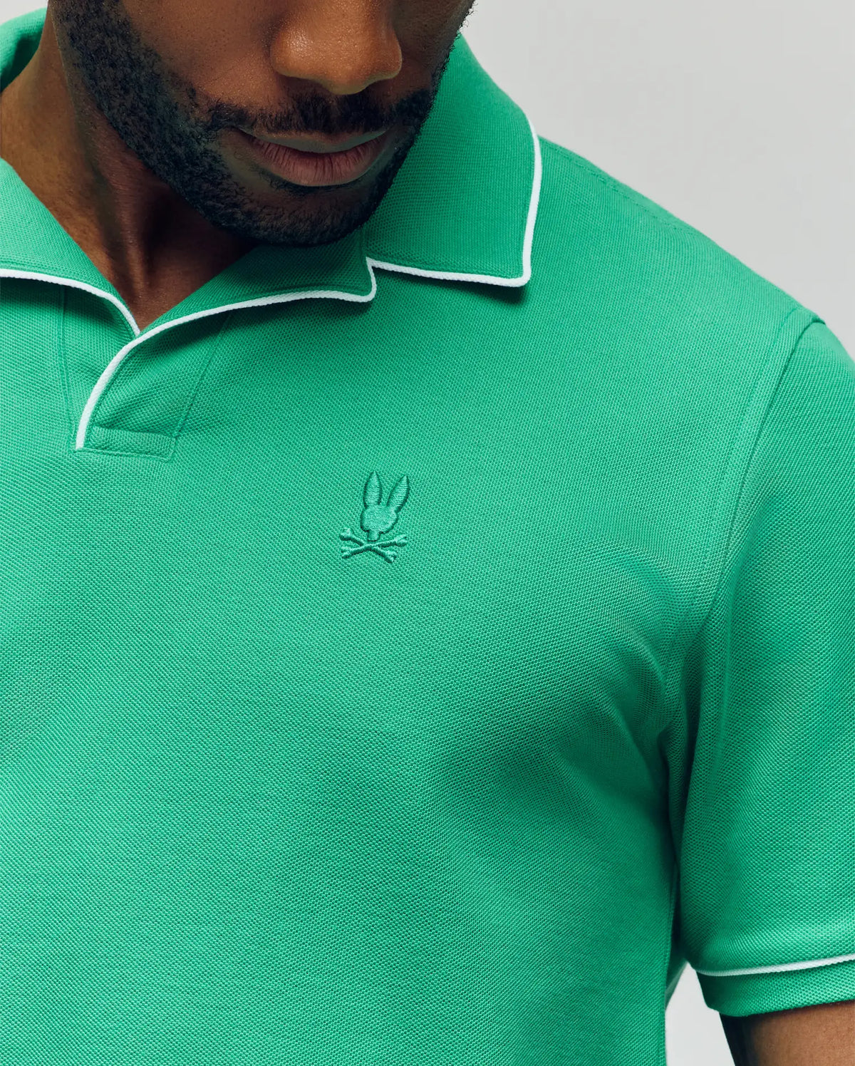 A man wears the Psycho Bunny MENS JAXON SOFT PIQUE CAMP COLLAR POLO (MT0100006) in green with white collar trim and an embroidered bunny logo, shown from lower face to upper torso.
