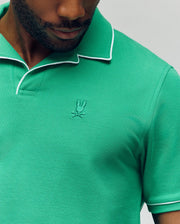 A man wears the Psycho Bunny MENS JAXON SOFT PIQUE CAMP COLLAR POLO (MT0100006) in green with white collar trim and an embroidered bunny logo, shown from lower face to upper torso.
