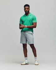 A man with short hair and a well-groomed beard poses confidently against a light gray background, wearing the Psycho Bunny MENS JAXON SOFT PIQUE CAMP COLLAR POLO (MT0100006) in green, paired with light gray shorts and white sneakers.