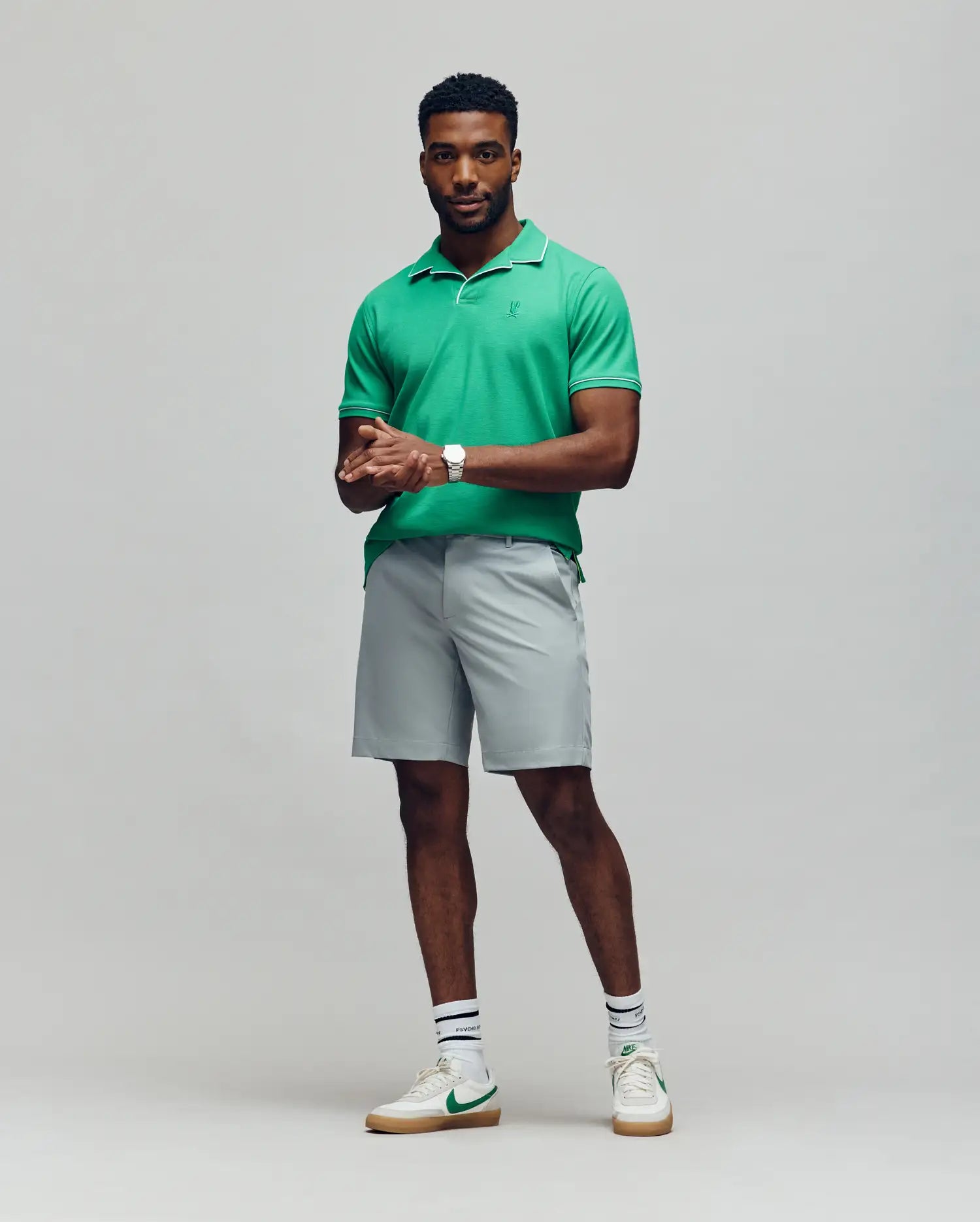 A man with short hair and a well-groomed beard poses confidently against a light gray background, wearing the Psycho Bunny MENS JAXON SOFT PIQUE CAMP COLLAR POLO (MT0100006) in green, paired with light gray shorts and white sneakers.