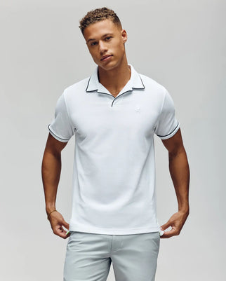 A man wearing the Psycho Bunny MENS JAXON SOFT PIQUE CAMP COLLAR POLO - MT0100006 in white with black trim and light gray pants stands against a plain background, looking at the camera with a neutral expression.
