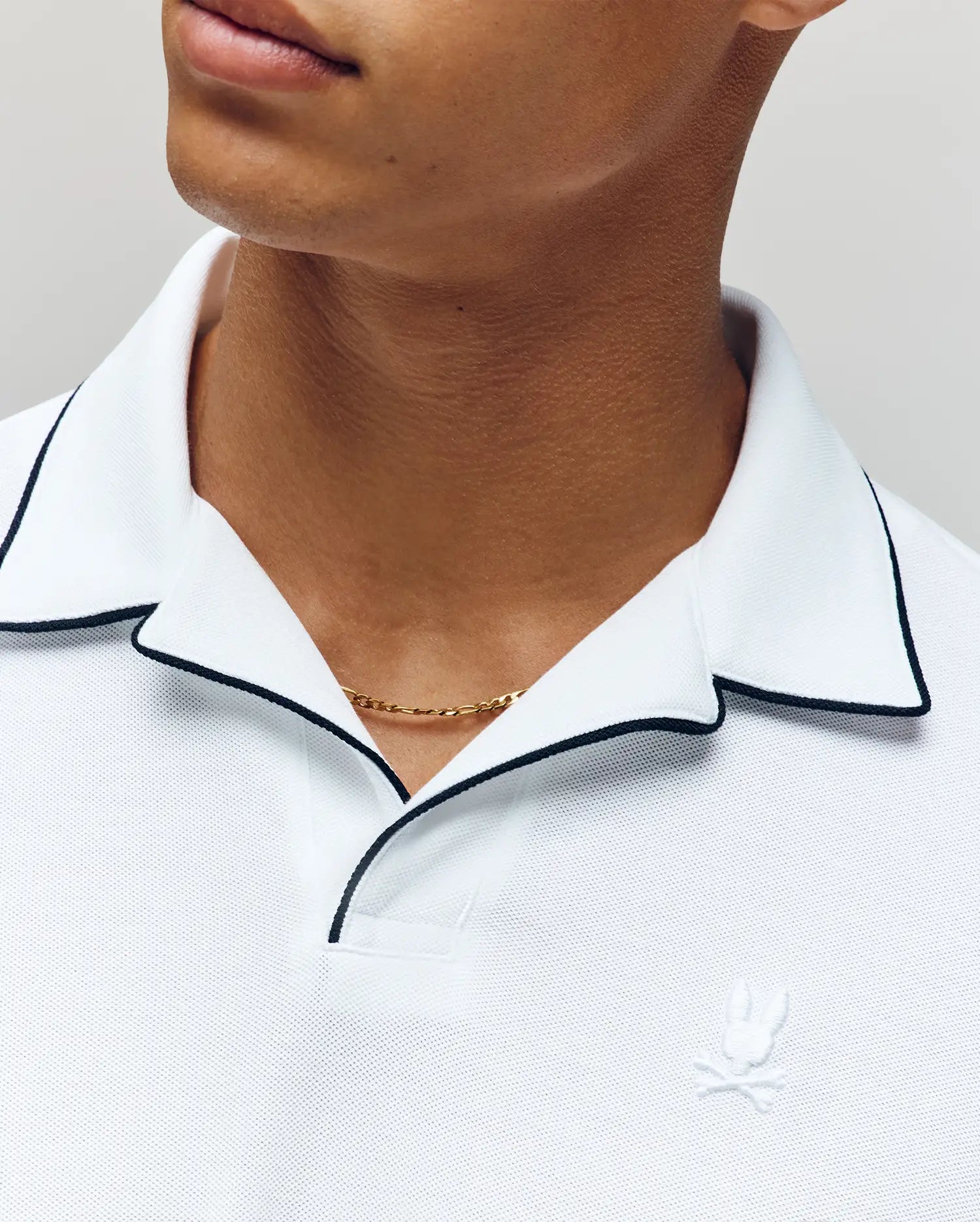 A man wears the Psycho Bunny MENS JAXON SOFT PIQUE CAMP COLLAR POLO (MT0100006), featuring black piping on the collar and a white embroidered rabbit with crossed bones logo, paired with a gold chain; only his neck and lower face are visible.