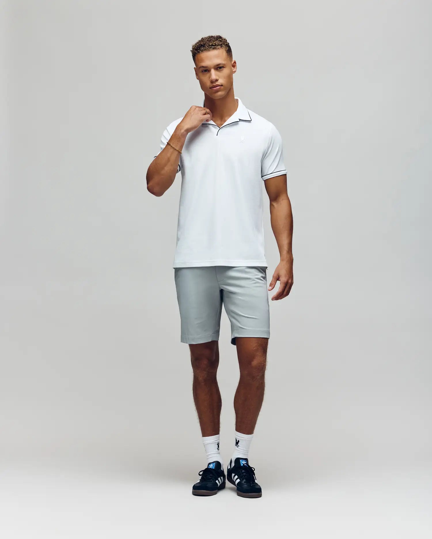 A man with short curly hair stands against a plain background, touching his neck and wearing a Psycho Bunny MENS JAXON SOFT PIQUE CAMP COLLAR POLO - MT0100006 in white, paired with light gray shorts, white socks, and black sneakers.