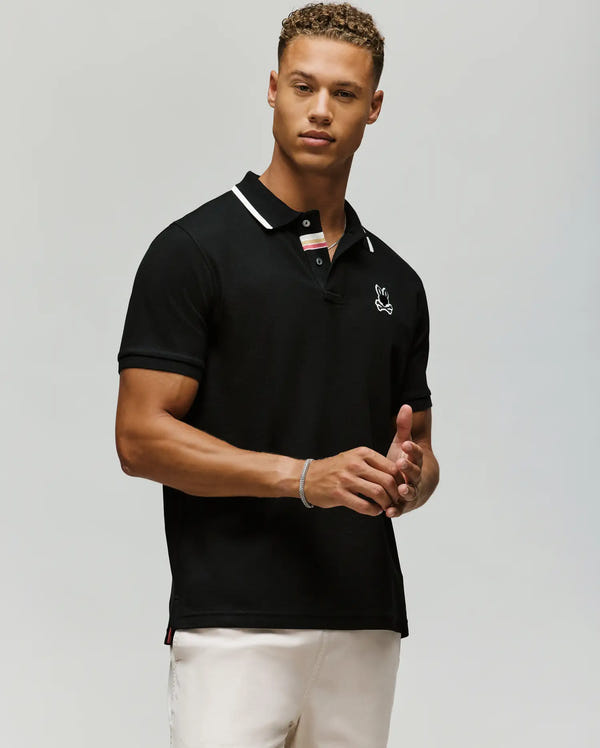 A young man with short curly hair wears Psycho Bunny’s MENS FENLEY PIQUE POLO (MT0100009) in black Pima cotton with white trim and an embroidered chest Bunny, paired with light pants against a plain light background.