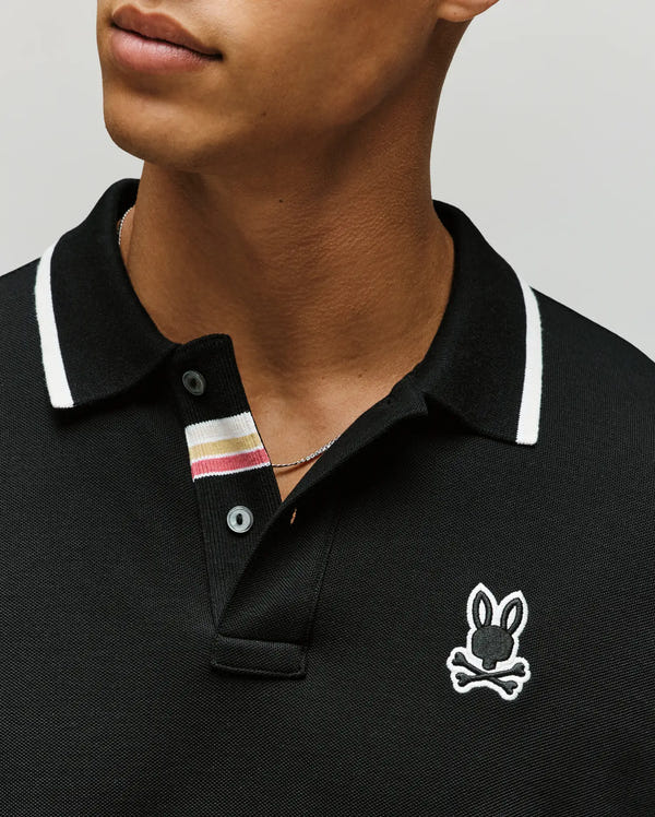 A man wears the Psycho Bunny MENS FENLEY PIQUE POLO (MT0100009) in black Pima cotton, with white-striped collar and embroidered Bunny and crossbones logo; only his lower face and upper torso are visible.