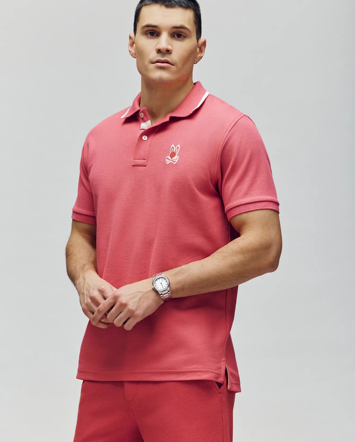 A man with short dark hair and a silver watch wears the Psycho Bunny MENS FENLEY PIQUE POLO - MT0100009 in coral pink, made from soft Pima cotton with an embroidered chest Bunny, plus matching shorts, standing against a light gray background.