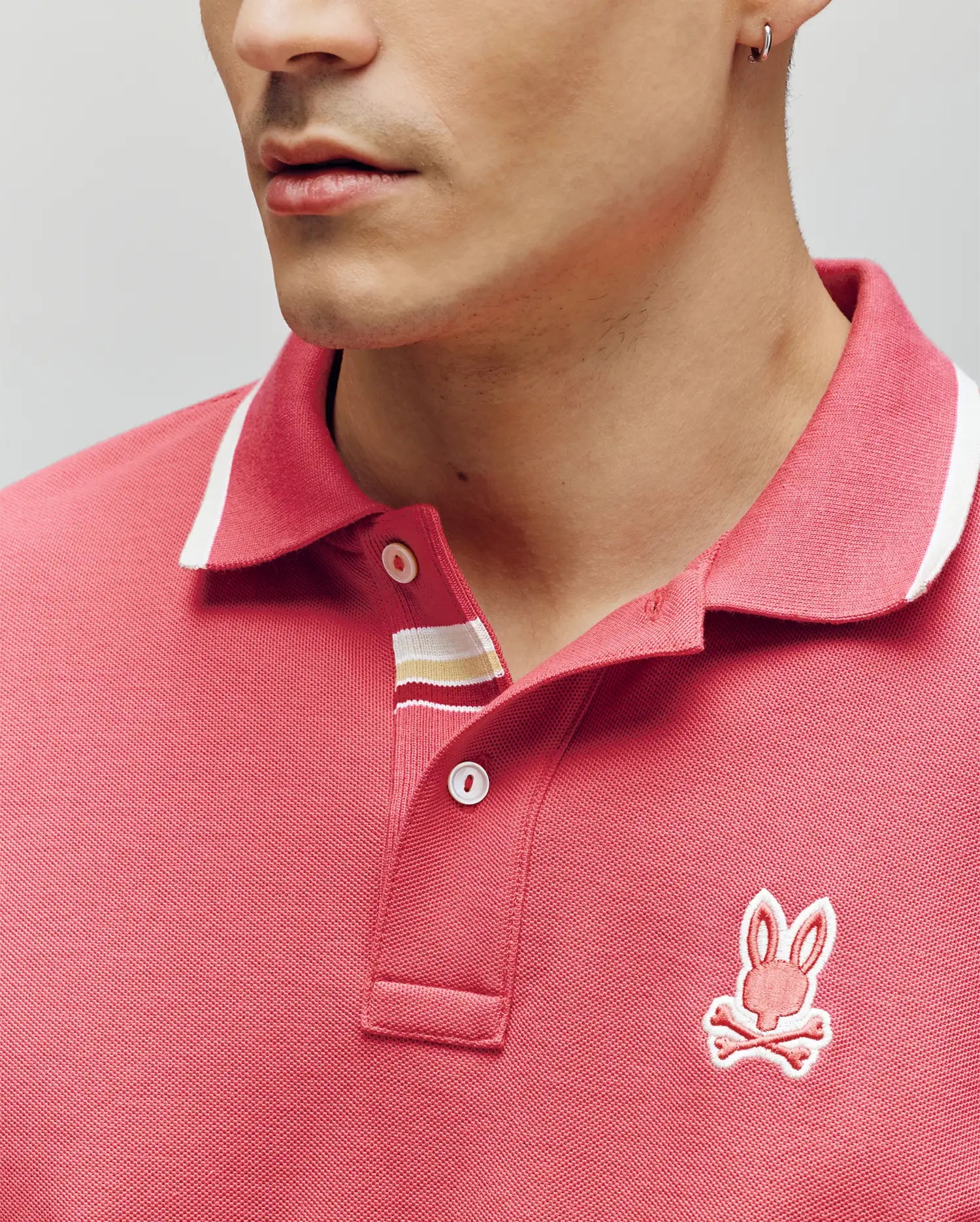 A man wears a red Psycho Bunny Mens Fenley Pique Polo (MT0100009) made from soft Pima cotton, with white trim and an embroidered Bunny and crossbones chest logo. Only his lower face and upper torso are visible.