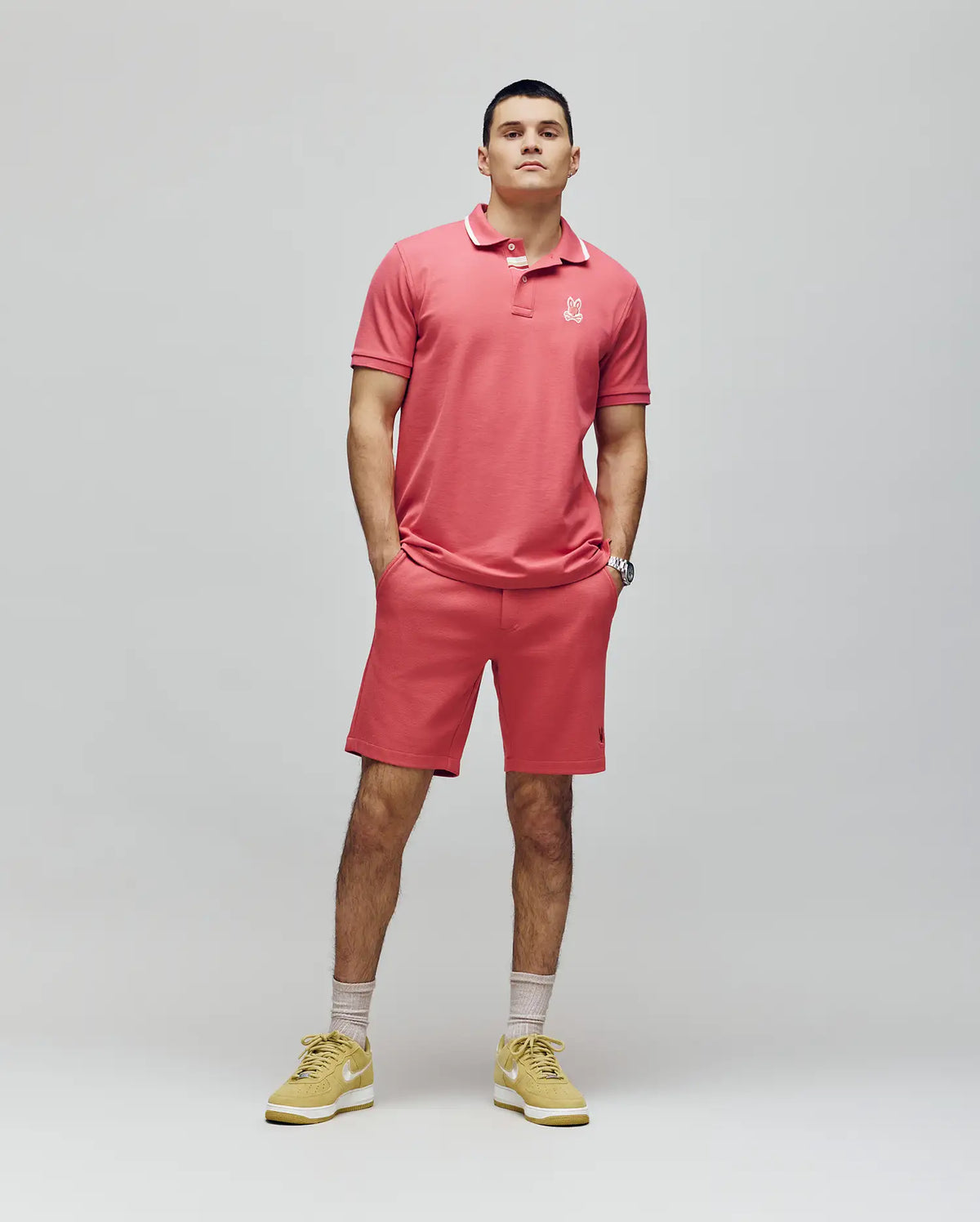 A man stands against a plain background wearing the Psycho Bunny MENS FENLEY PIQUE POLO - MT0100009 in pink, with an embroidered chest Bunny, matching shorts, yellow sneakers, and light-colored socks. His hands are in his pockets as he looks ahead neutrally.