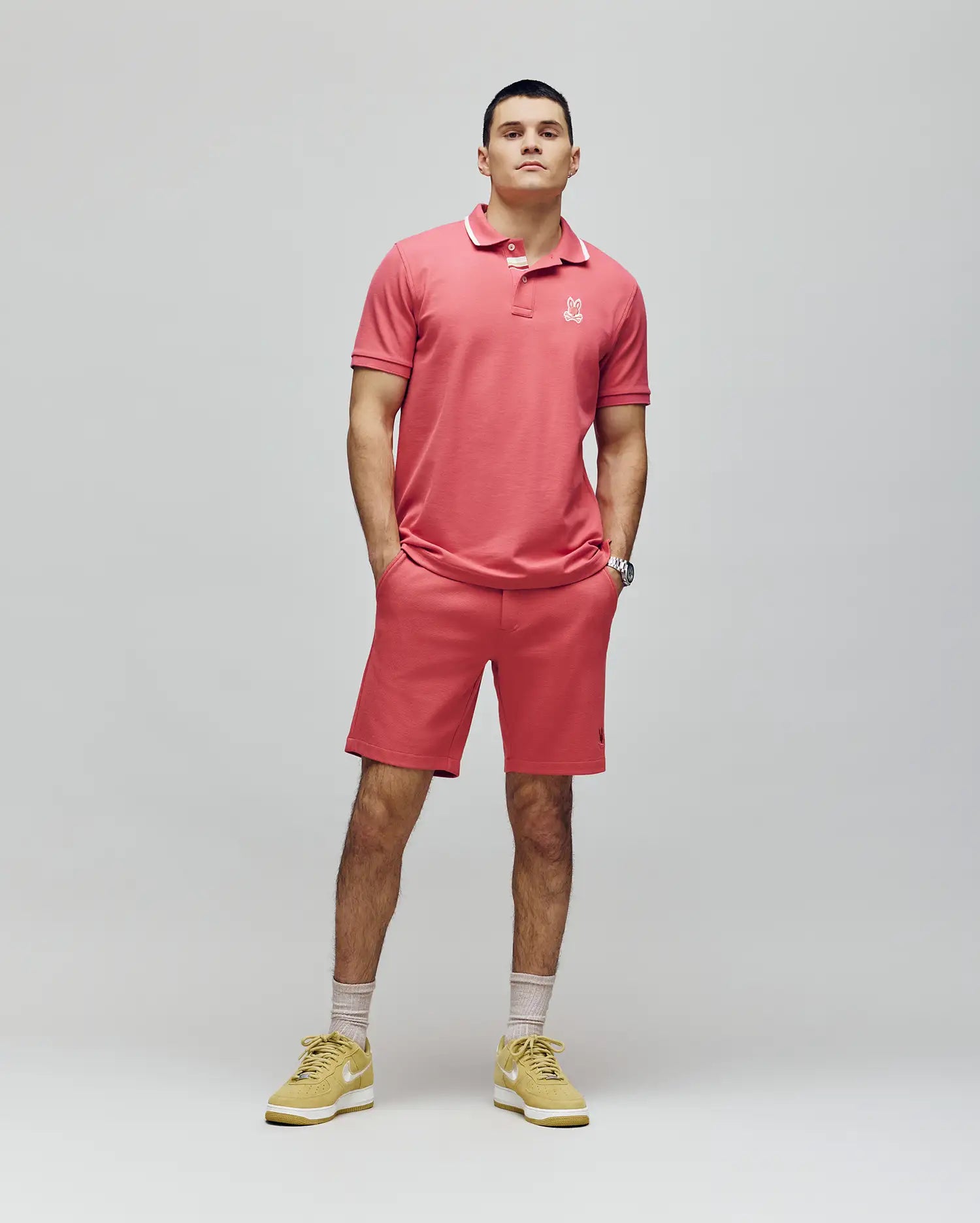 A man stands against a plain background wearing the Psycho Bunny MENS FENLEY PIQUE POLO - MT0100009 in pink, with an embroidered chest Bunny, matching shorts, yellow sneakers, and light-colored socks. His hands are in his pockets as he looks ahead neutrally.