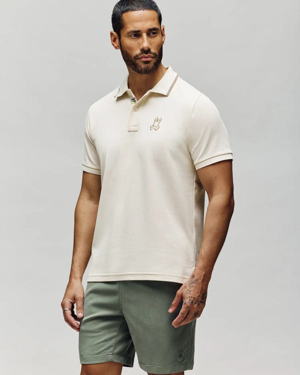 A man with short dark hair and a beard wears the Psycho Bunny MENS FENLEY PIQUE POLO - MT0100009 in cream, featuring an embroidered chest logo, paired with olive green shorts, standing against a plain light gray background.