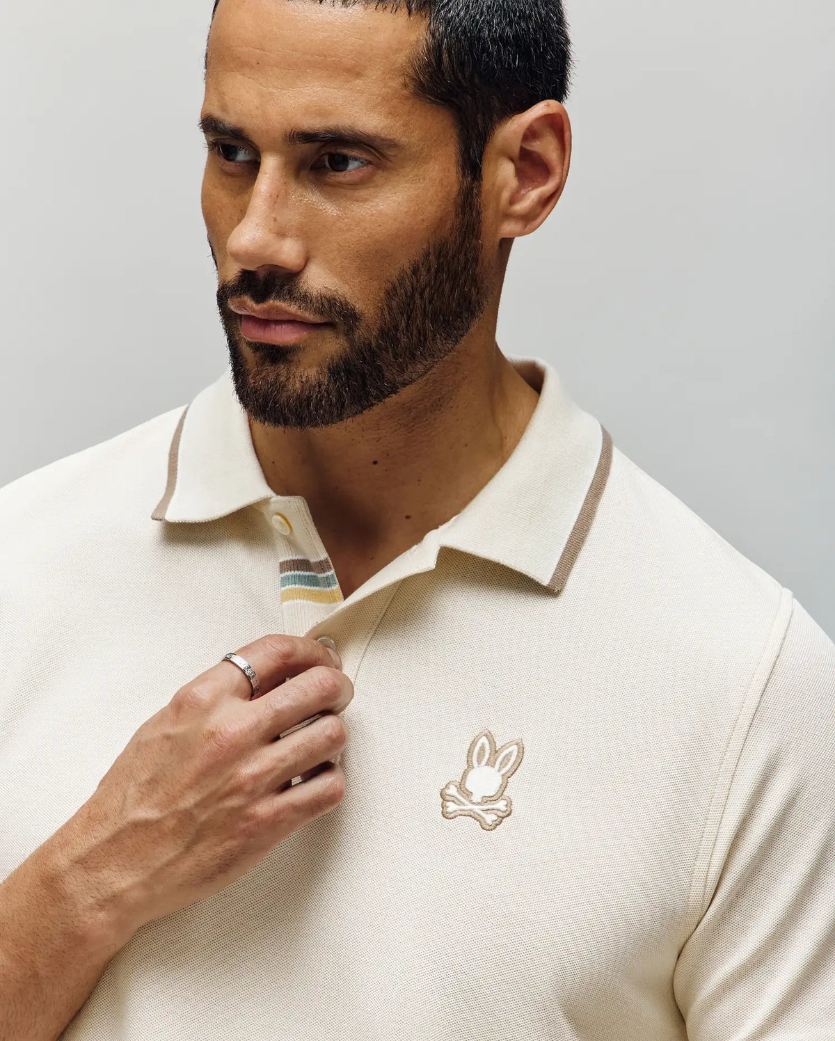 A man with short dark hair and a beard wears the Psycho Bunny MEN'S FENLEY PIQUE POLO (MT0100009) in cream, featuring Mother-of-Pearl buttons and an embroidered bunny logo. He looks thoughtfully to the side, a ring on his finger.