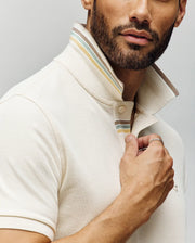 A bearded man wears the Psycho Bunny MENS FENLEY PIQUE POLO (MT0100009) in cream Pima cotton with the collar popped, showing striped detail inside. He holds the collar, highlighting Mother-of-Pearl buttons and a ring on his finger.
