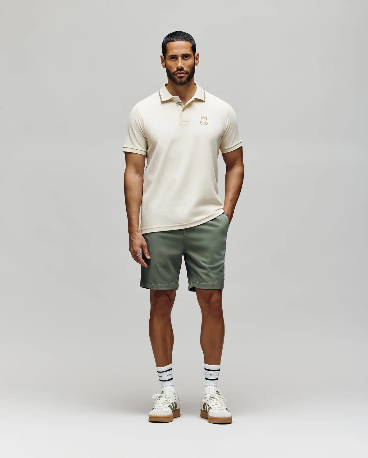 A man faces forward wearing the Psycho Bunny Mens Fenley Pique Polo (MT0100009), sage green shorts, white sneakers with gum soles, and white socks with navy stripes, against a plain light grey background.