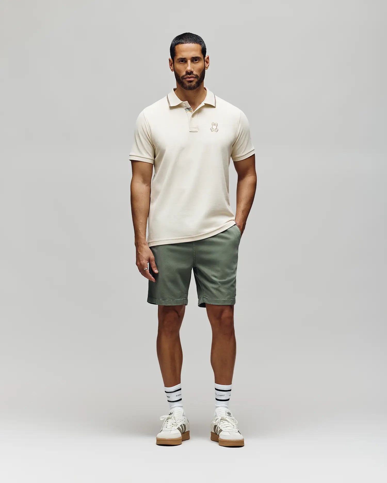A man faces forward wearing the Psycho Bunny Mens Fenley Pique Polo (MT0100009), sage green shorts, white sneakers with gum soles, and white socks with navy stripes, against a plain light grey background.