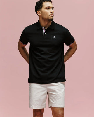 A man stands against a pink background, wearing the Psycho Bunny MENS ARCHIVE CLASSIC BUNNY PIQUE POLO (MT0100010) in black with an embroidered logo on the chest and light shorts. His arms are behind his back as he gazes to the side.