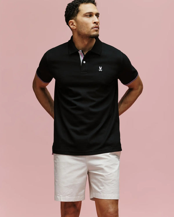 A man stands against a pink background, wearing the Psycho Bunny MENS ARCHIVE CLASSIC BUNNY PIQUE POLO (MT0100010) in black with an embroidered logo on the chest and light shorts. His arms are behind his back as he gazes to the side.