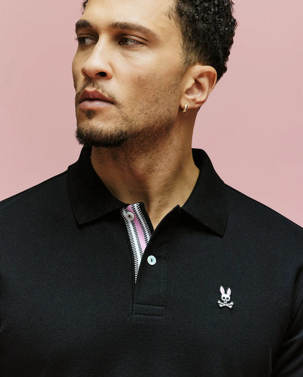 A man with short curly hair and a gold hoop earring looks to the side, wearing a black Psycho Bunny MENS ARCHIVE CLASSIC BUNNY PIQUE POLO (MT0100010) with an embroidered bunny logo, set against a pink background.