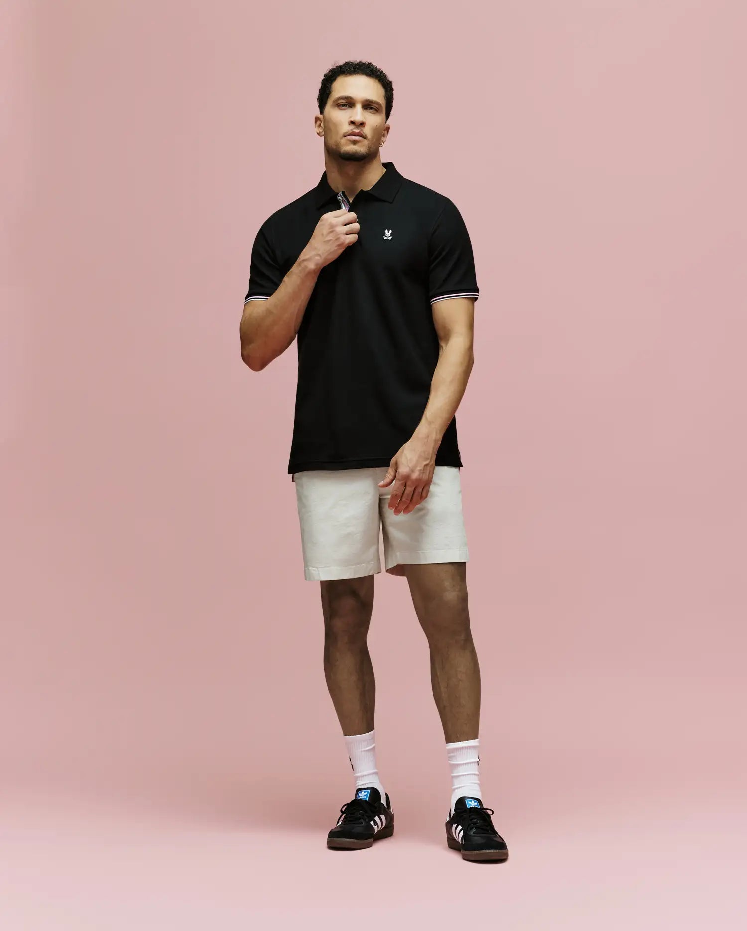 A man stands against a pink background wearing Psycho Bunny’s MENS ARCHIVE CLASSIC BUNNY PIQUE POLO - MT0100010, light beige shorts, white socks, and black Adidas sneakers. He holds the polo’s collar with one hand and looks forward.