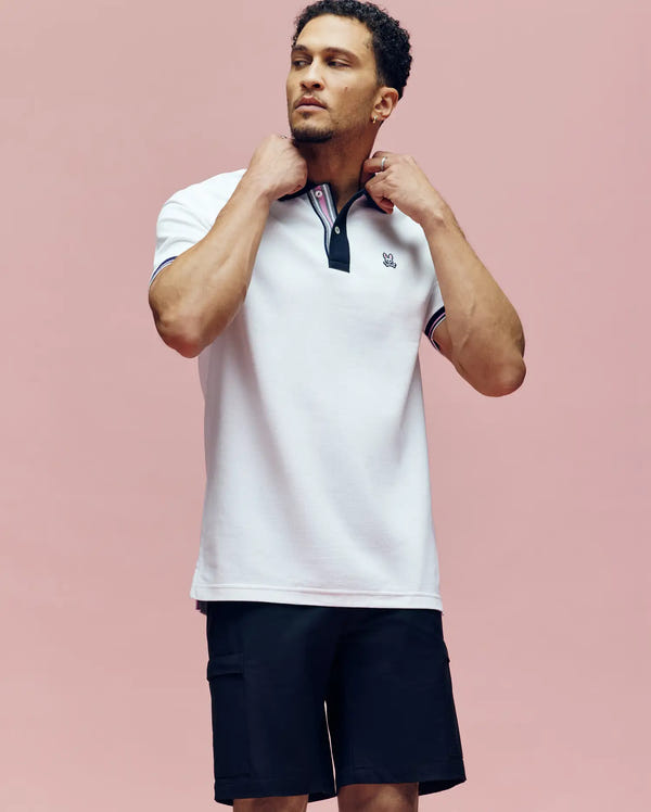 A man stands against a pink background, adjusting the collar of his white Psycho Bunny MENS ARCHIVE CLASSIC BUNNY PIQUE POLO (MT0100010) with dark trim. He is also wearing black shorts and gazing slightly upwards.