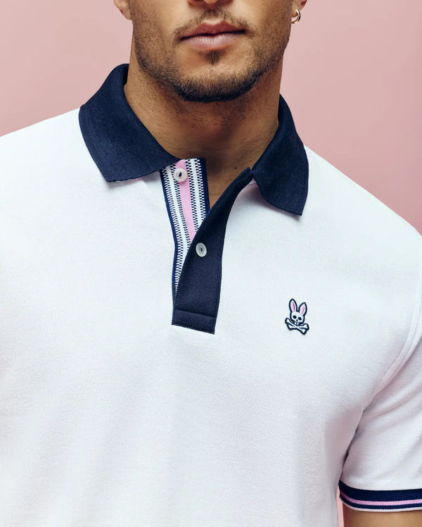 A man wears the Psycho Bunny MENS ARCHIVE CLASSIC BUNNY PIQUE POLO (MT0100010), a white Pima cotton shirt with a black collar, striped placket, and embroidered Bunny skull logo, standing against a pink background.