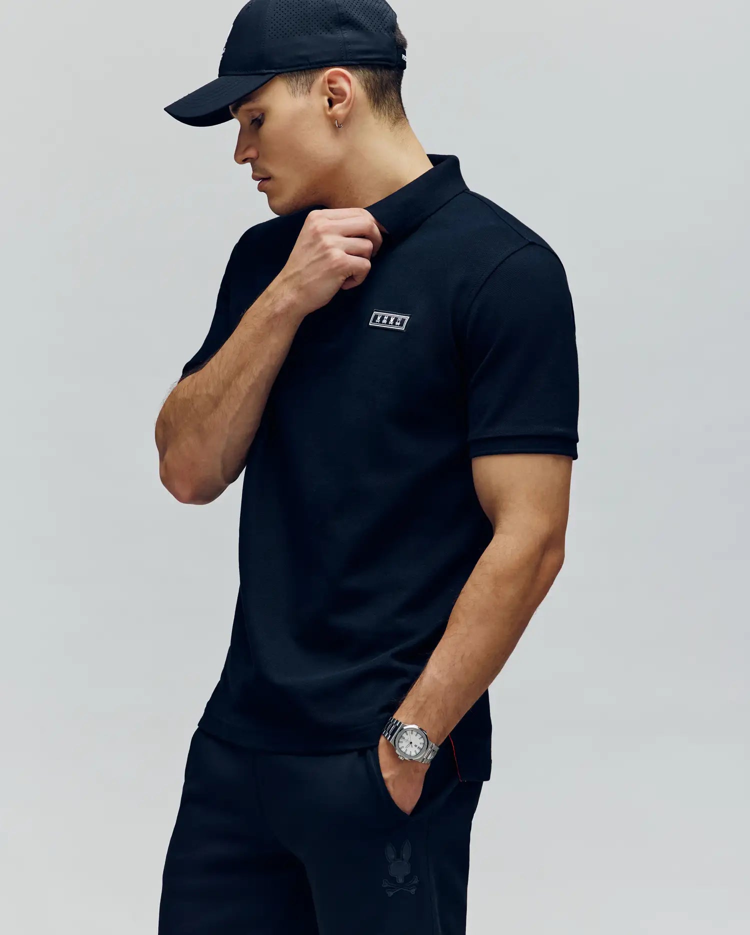A man in a Psycho Bunny MENS AXTON PIQUE POLO (MT0100011), black cap, and pants stands against a plain background, wearing a watch on his left wrist and touching his collar with his right hand while looking to the side.