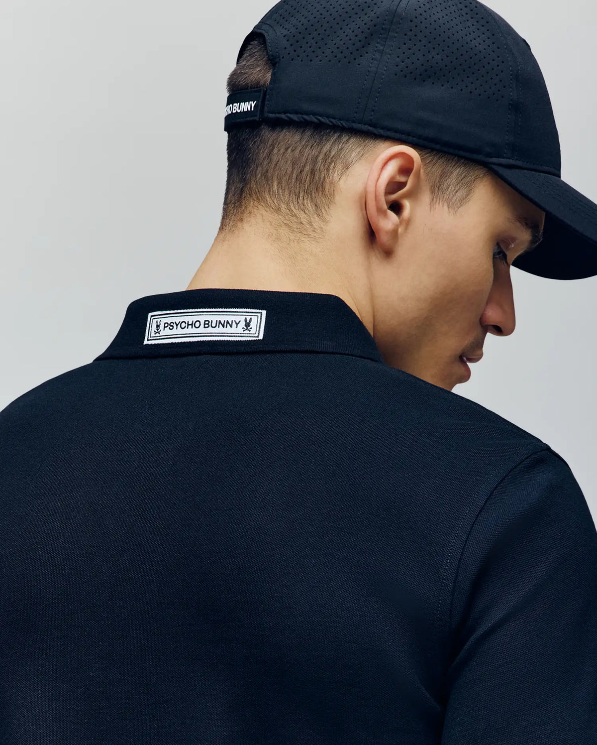 A person is seen from behind, slightly turned, wearing a black Psycho Bunny MENS AXTON PIQUE POLO - MT0100011 and a black cap, with the white "Psycho Bunny" label visible on the back of the collar.