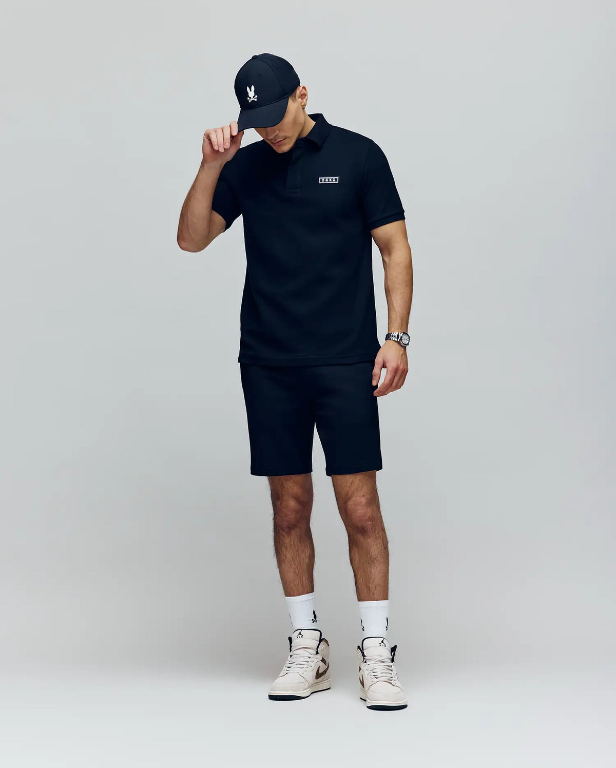 A man in a Psycho Bunny MENS AXTON PIQUE POLO (MT0100011), black shorts, a black cap, and white high-top sneakers stands against a plain light background, tilting his cap downward with one hand.