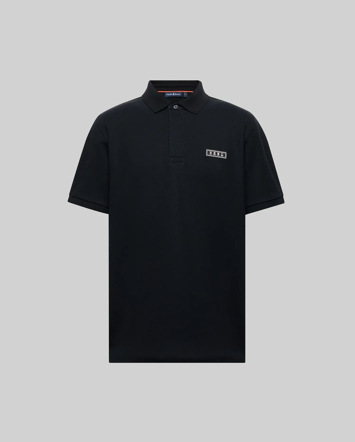 The Psycho Bunny MENS AXTON PIQUE POLO (MT0100011) is a plain black Pima cotton short-sleeve polo with a collar, three buttons, and a jacquard-woven logo patch on the left chest, shown against a light gray background.