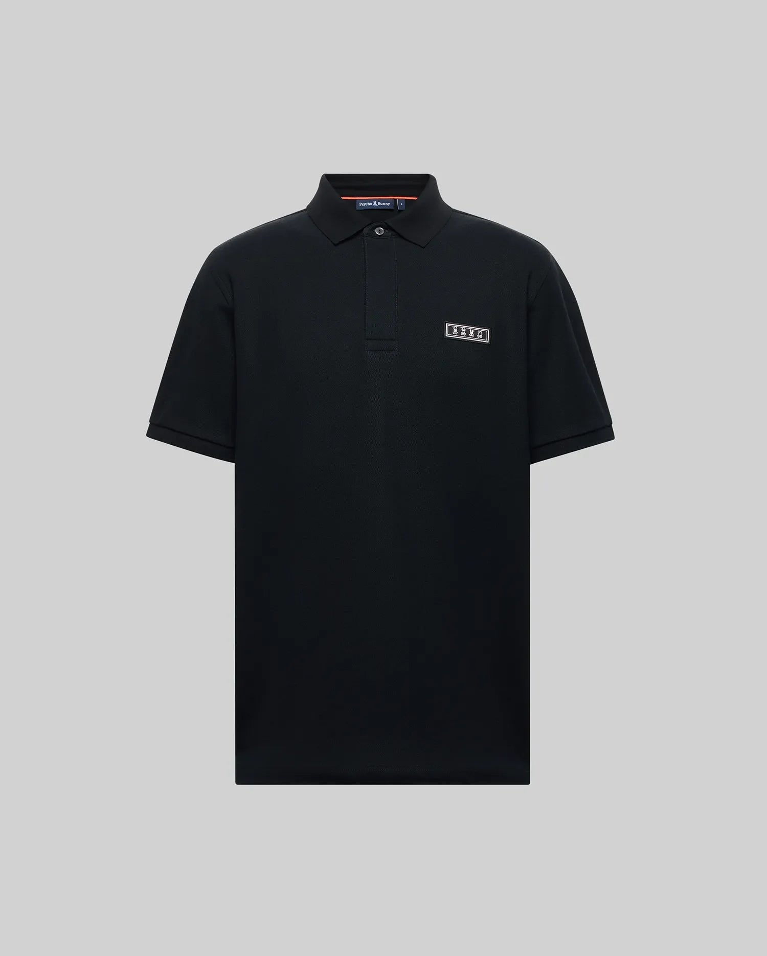 The Psycho Bunny MENS AXTON PIQUE POLO (MT0100011) is a plain black Pima cotton short-sleeve polo with a collar, three buttons, and a jacquard-woven logo patch on the left chest, shown against a light gray background.