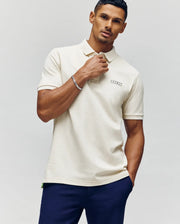A man with short hair and a trimmed beard wears the cream Psycho Bunny MENS AXTON PIQUE POLO (MT0100011) and dark blue pants, standing against a plain light background, one hand touching his collar.